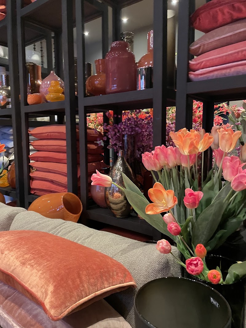 Binnen in de winkel: zijden tulpen, fluwelen kussens in koraal en roze, vazen en woonaccessoires op zwarte stellingen.